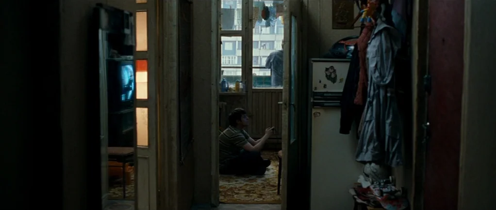 Elena (2011) movie still: medium — A cluttered apartment interior with a long corridor leading toward an open balcony window. A teenage boy sits on…