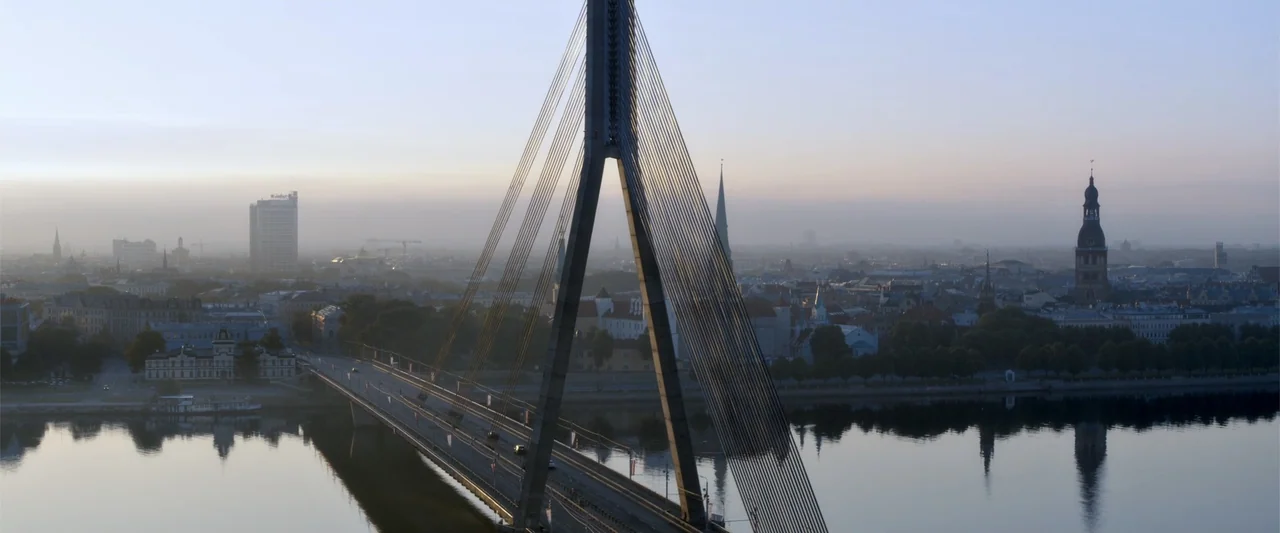 The Good Neighbor (2022) movie still: establishing — A cable-stayed bridge crosses a wide river with a sprawling historic city in the background.…