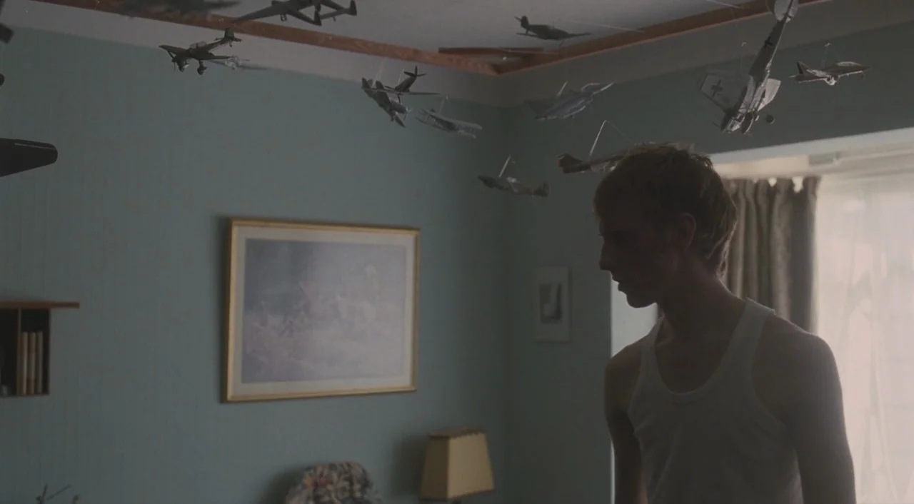 Broken (2012) movie still: medium — A room with blue-grey walls features many model airplanes suspended from the ceiling. A young man in a white…