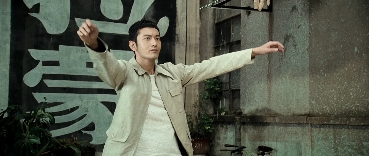 Ip Man 2 (2010) movie still: medium — A weathered outdoor wall with faded painted calligraphy stands behind a man exercising. The man in a beige…