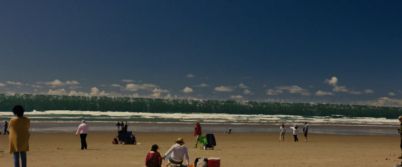 Knock at the Cabin (2023) movie still: wide — A wide beach under a deep blue sky with a massive breaking wave. Several people are scattered across…