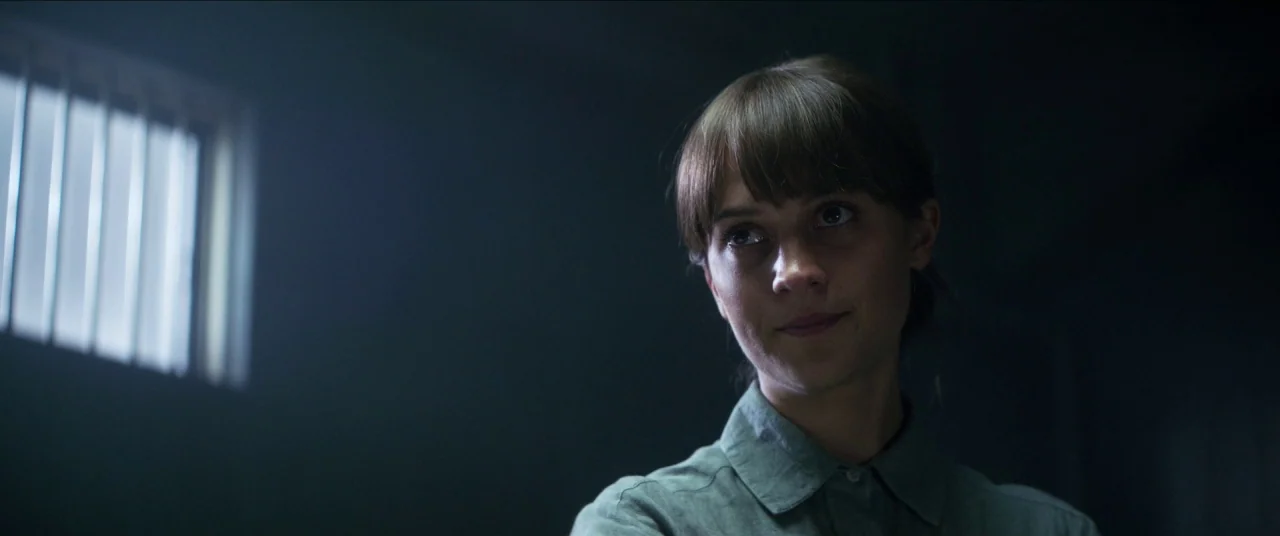 Earthquake Bird movie still: medium — A dark, spartan room features a single barred window letting in slivers of daylight. A young woman with brown…