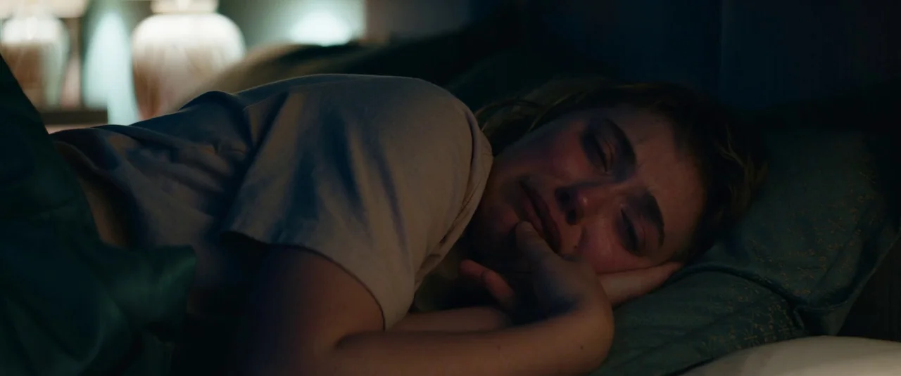Vivarium (2019) movie still: closeup — A dimly lit room with blue patterned bedding and a glowing lamp base. A young woman lies on her side with her…