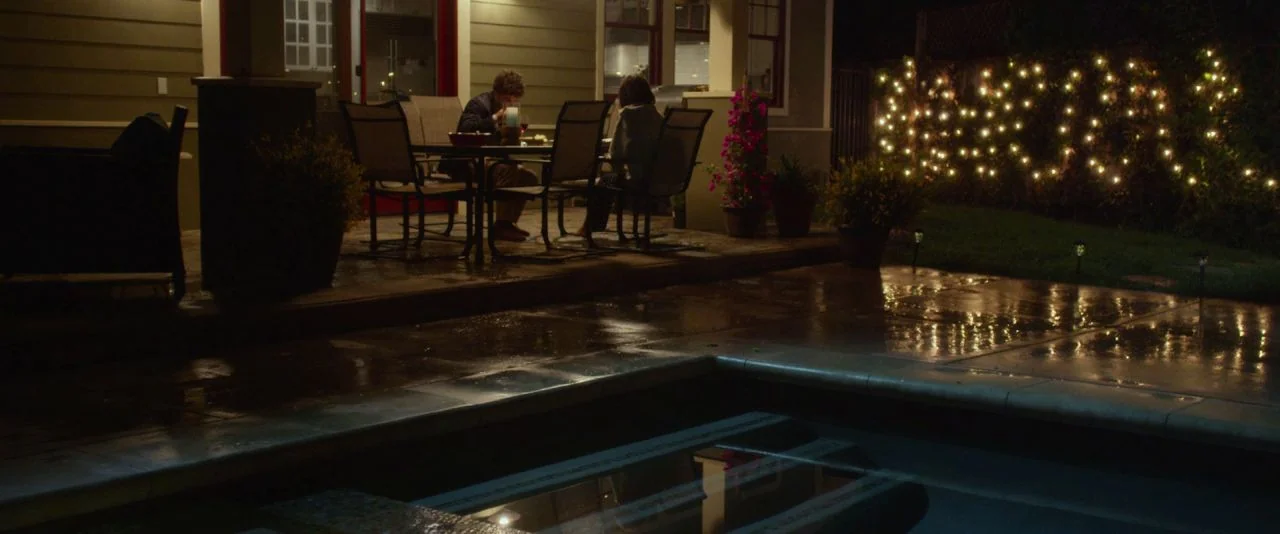 Night Owls (2015) movie still: wide — A residential backyard patio at night featuring a dining table near a dark house. A couple sits at the table…