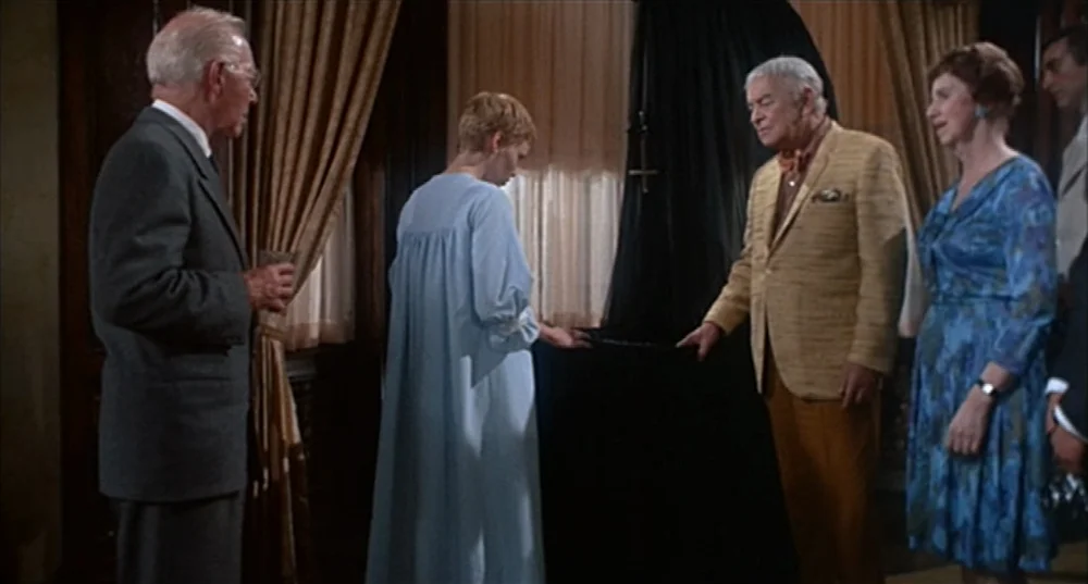Rosemary’s Baby (1968) movie still: medium — A dimly lit room with heavy curtains and wooden wall paneling background. A woman in a light blue gown…