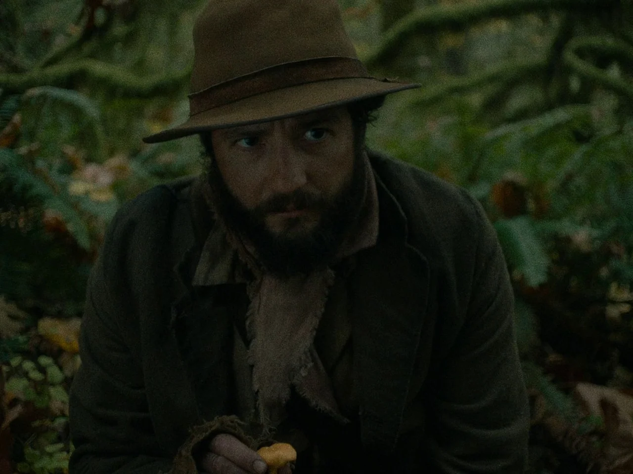 First Cow (2019) movie still: medium — An overgrown woods floor covered in ferns and mossy branches in the Pacific Northwest. A man wearing a rustic…