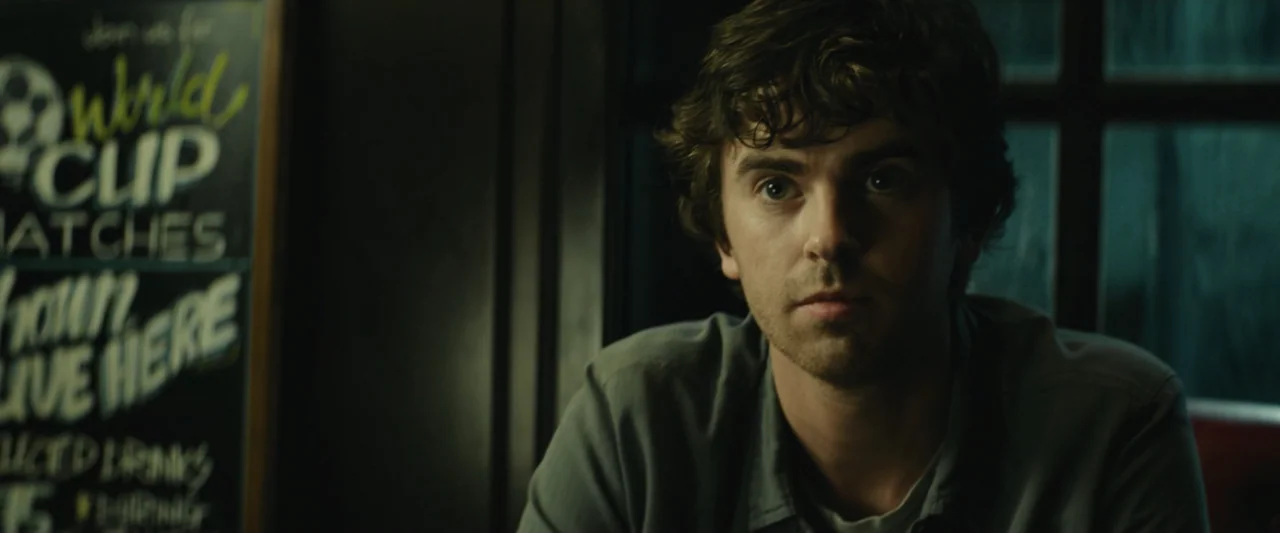 The Vault (2021) movie still: medium — A dimly lit pub interior features a dark wooden wall and a chalkboard menu. A man in his twenties with brown…
