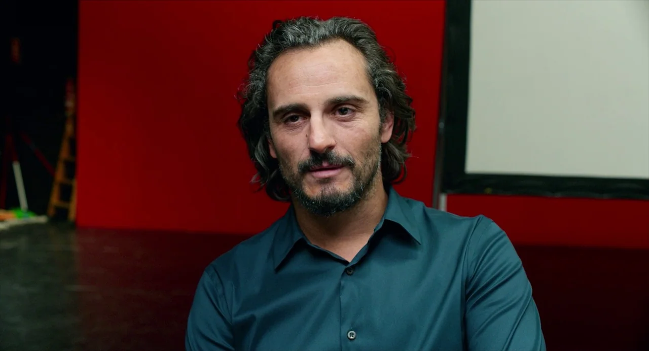 Pain and Glory (2019) movie still: medium — A man stands in a studio space with a bright red backdrop and white projection screen. The space has…