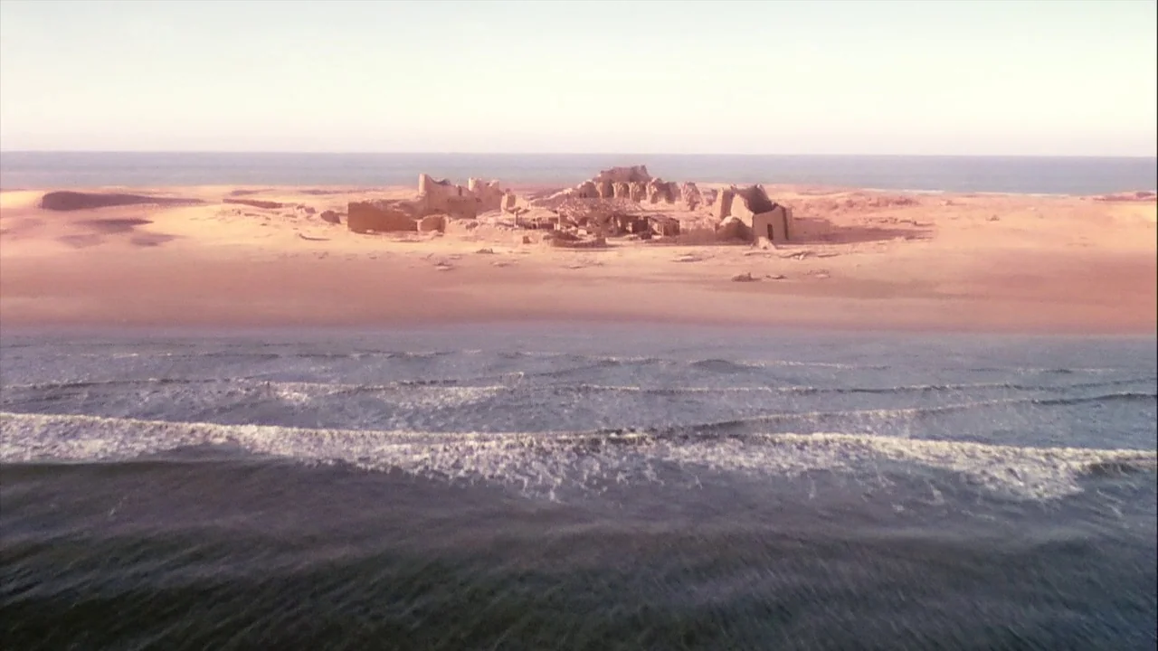The Manchurian Candidate (2004) movie still: establishing — Stone ruins stand isolated on a sandy shoreline beside a calm ocean. Waves gently…