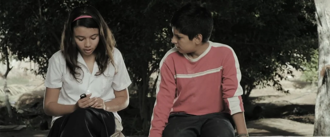 At Dawn (2013) movie still: medium — Two teenagers sit outdoors in front of a backdrop of dense trees and foliage. The girl holds a small object in…