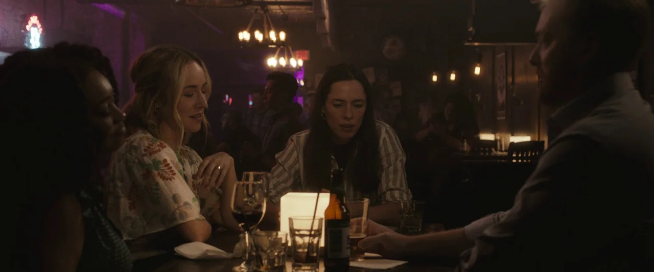 The Night House (2020) movie still: medium — A dimly lit bar interior featuring a wooden table occupied by three people. The group interacts while…
