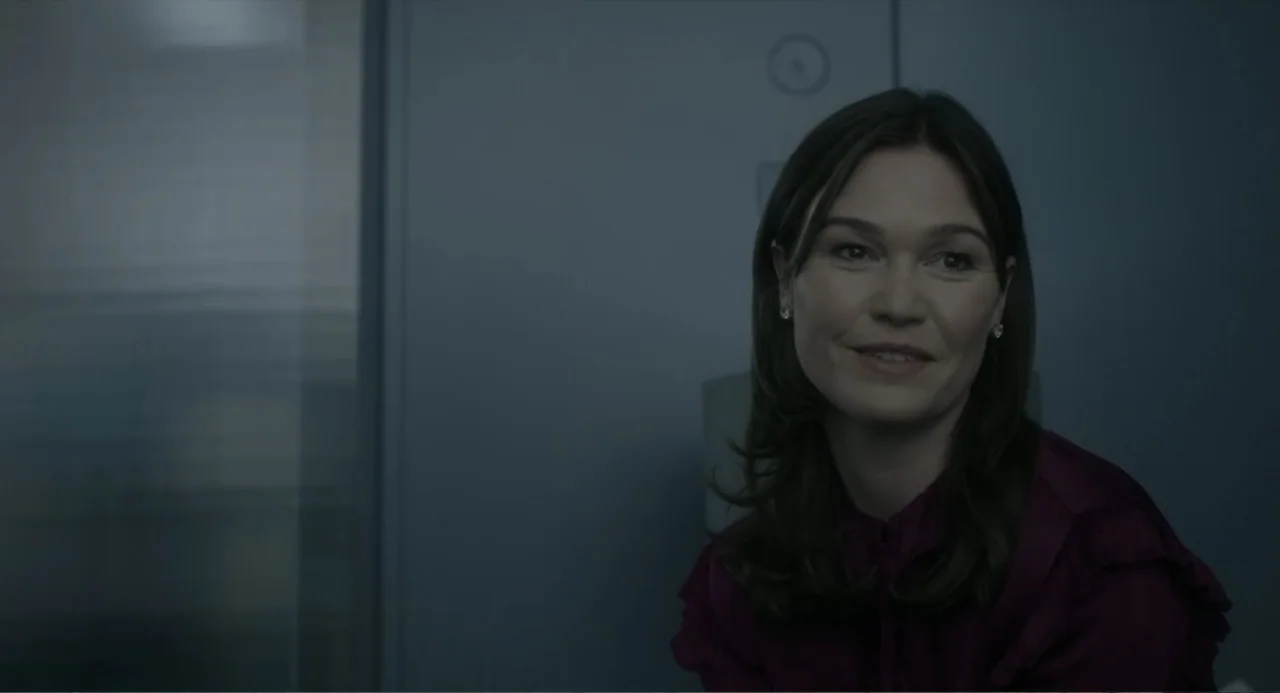 Orphan: First Kill (2022) movie still: medium — An office interior features a grey wall and a dark, blurred reflective surface behind. A woman in a…