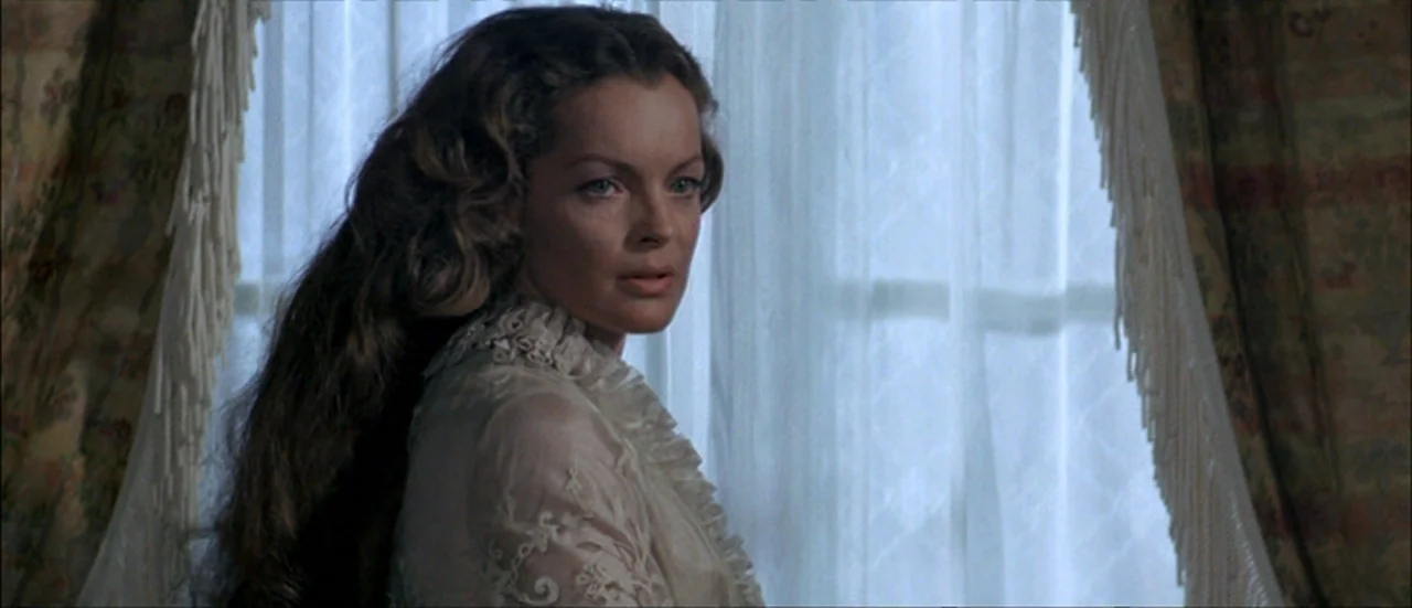 Ludwig (1973) movie still: closeup — A woman stands indoors in front of translucent white curtains with decorative fringe trim. She looks off-screen…