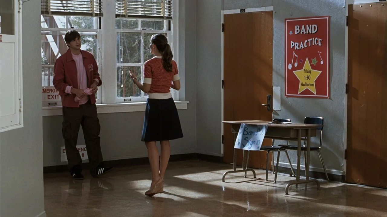 F Valentine's Day (2026) movie still: fullbody — A school hallway features gray walls, wooden doors, and a Band Practice flyer. A man and a woman…