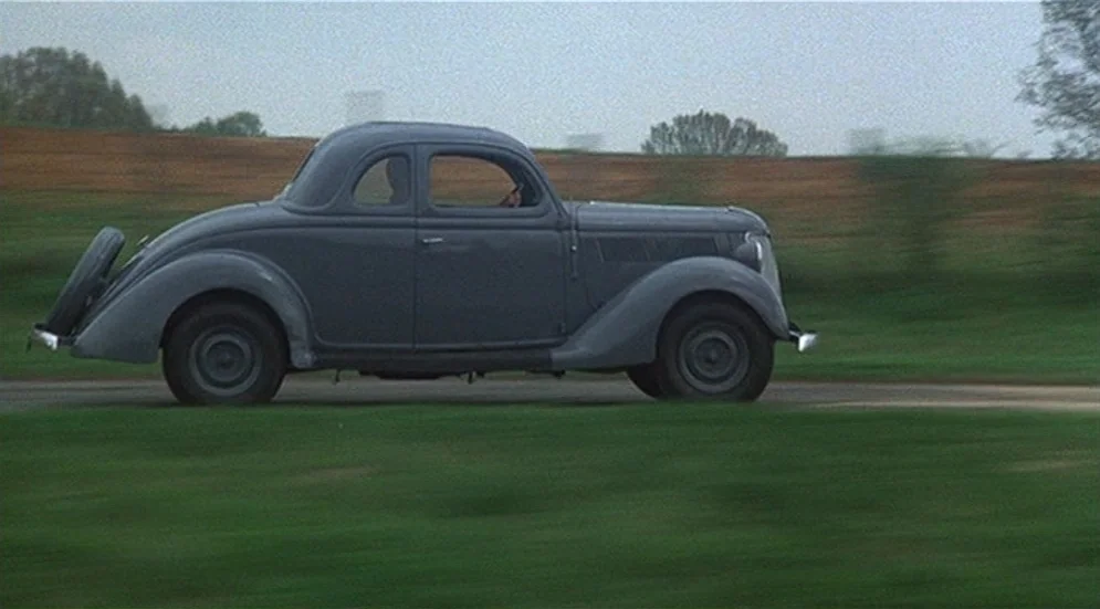 Thieves Like Us (1974) movie still: fullbody — A vintage 1930s grey coupe drives along a blurred country road. A person drives the vehicle across a…