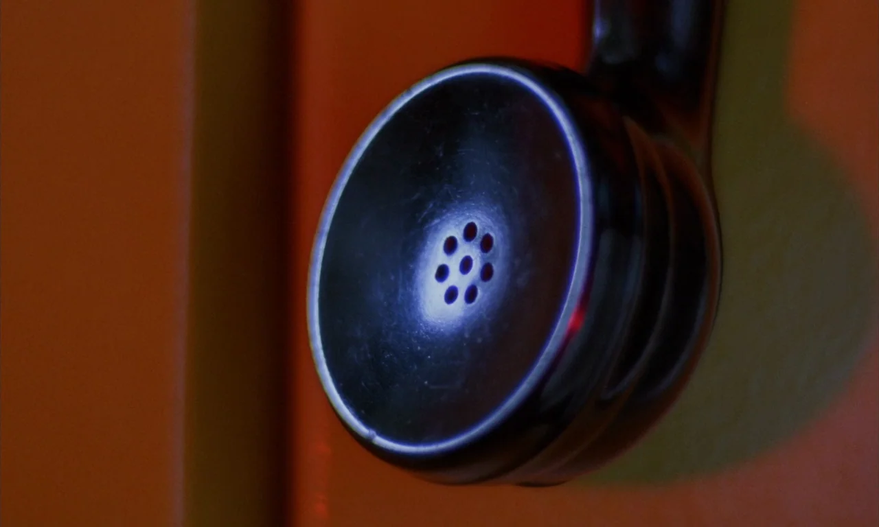 The Church (1989) movie still: closeup — A close-up of a black telephone receiver positioned against a vibrant orange vertical surface. The receiver…
