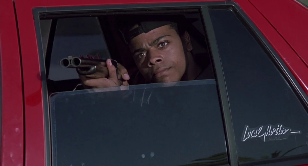 Boyz In The Hood (1991) movie still: medium — The interior of a red vehicle is visible through the open side window frame. A young man holds a…