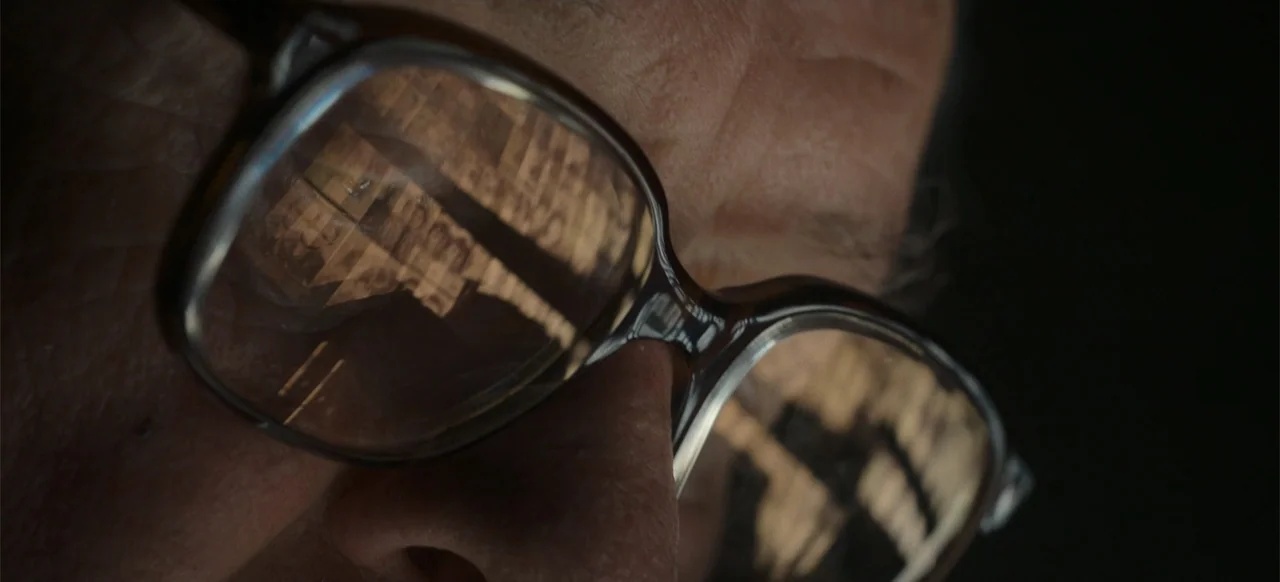 One Life (2023) movie still: closeup — An extreme close-up focuses on a person's skin and eyeglasses. The reflection of a building structure is…