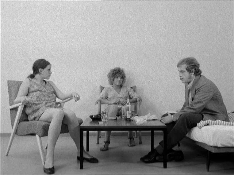 Katzelmacher (1969) movie still: fullbody — A plain white room with three people seated around a low black coffee table. Two women sit in chairs…