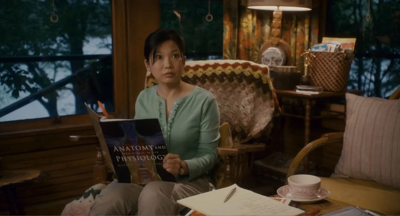 Grown Ups (2010) movie still: medium — A cozy rustic room features large windows and wooden wall paneling with cluttered decor. A woman sits in a…