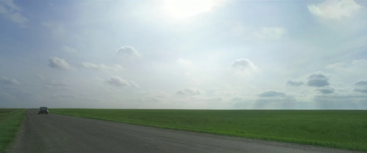 In The Tall Grass (2019) movie still: establishing — A flat, grassy horizon stretches out under a vast sky filled with scattered clouds. An asphalt…