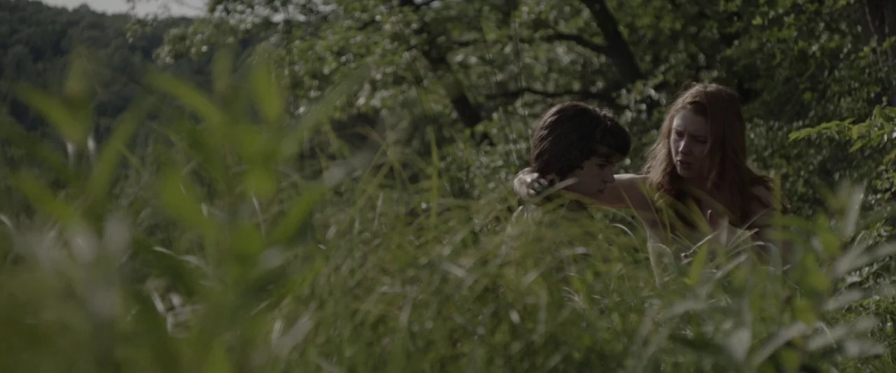 Here Alone movie still: medium — A dense patch of tall grass and trees forms a wooded natural clearing. A young woman and a young boy sit together…
