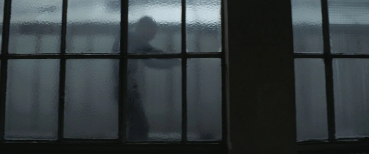 Colt 45 (2014) movie still: medium — A man stands behind large industrial windows with textured, frosted glass panes. He faces sideways, his form…