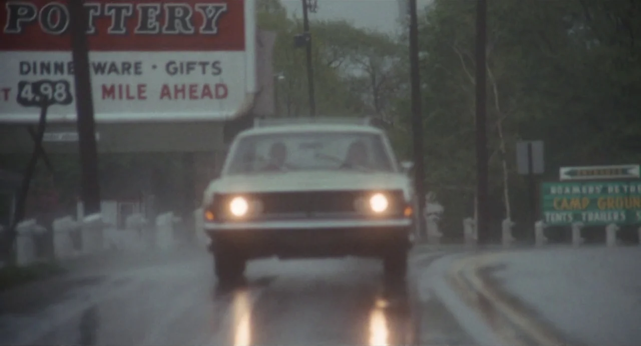 The Rain People (1969) movie still: wide — A wet asphalt road passes a large billboard advertising pottery and a campground entrance. A white sedan…