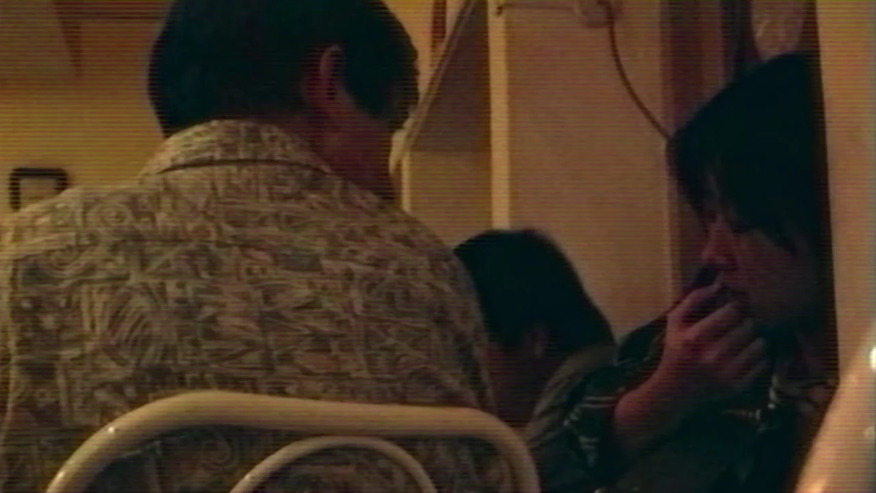 Shirkers (2018) movie still: medium — A dimly lit indoor booth setting with a patterned light shirt visible in the foreground. A woman sits across…
