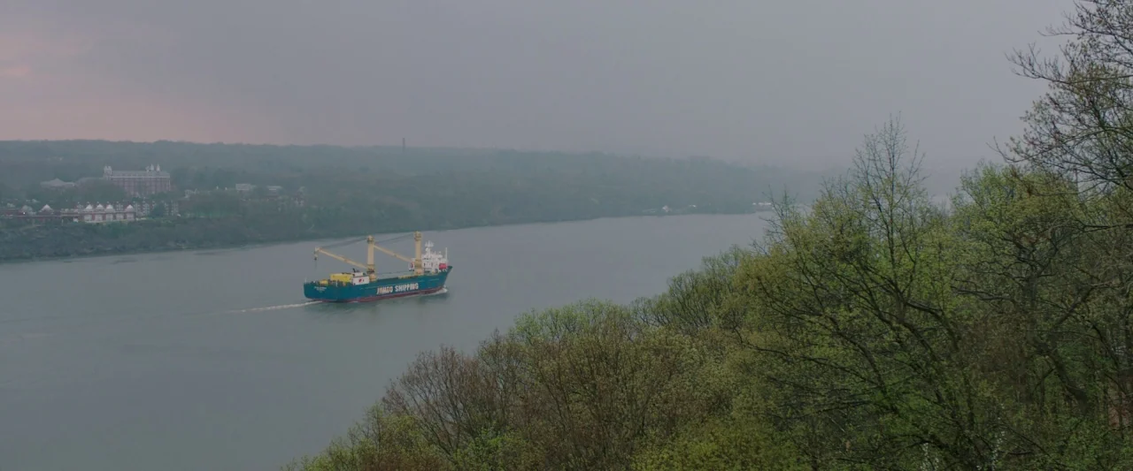 Swallow (2020) movie still: establishing — A wide river flows through a landscape under heavy gray overcast clouds. A cargo ship with two cranes…