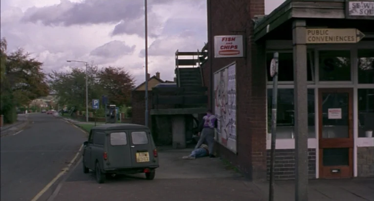 A Room For Romeo Brass (1999) movie still: wide — An overcast day outside a brick commercial building with fish and chips signage. A figure stands…