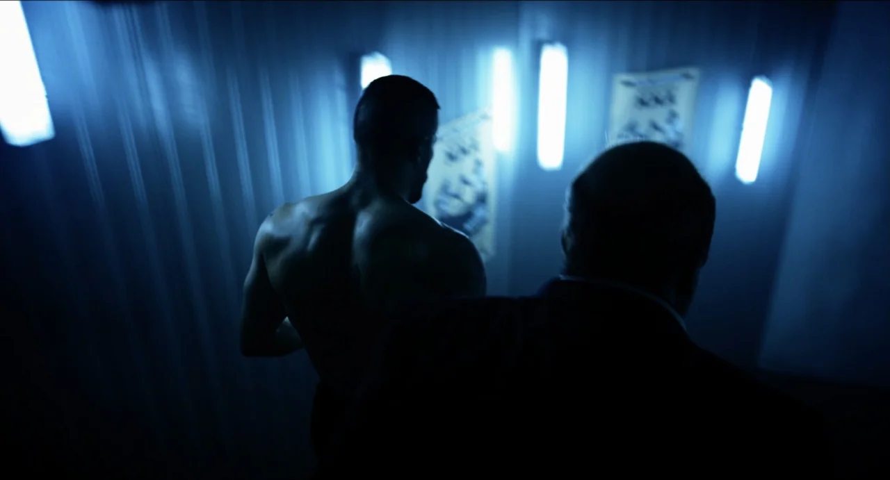 Boyka: Undisputed IV (2016) movie still: medium — A dark, narrow corridor features blue fluorescent lighting fixtures mounted on the walls. Two men…