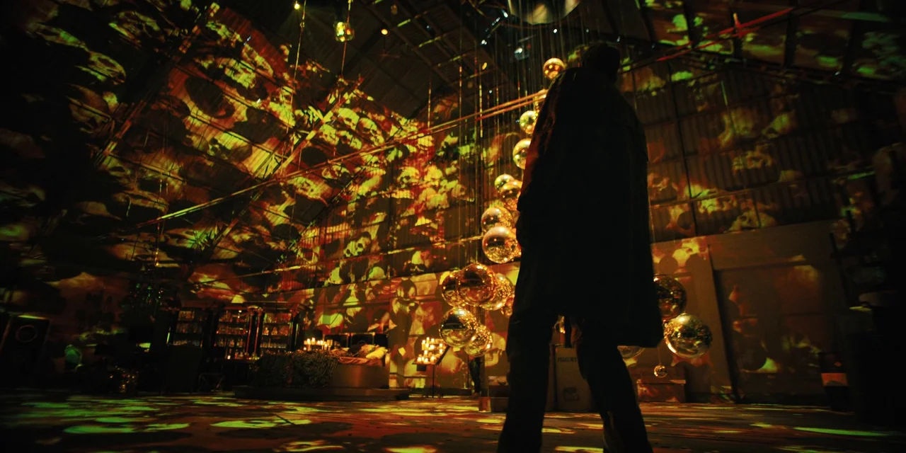 Silent Night (2023) movie still: fullbody — A dark warehouse interior contains a cluster of suspended metallic spheres and projections. A man stands…