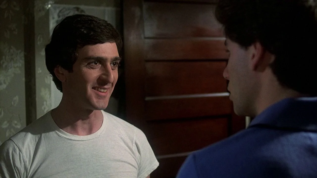Saturday Night Fever (1977) movie still: medium — A dimly lit home interior features floral wallpaper and a wooden door. A man in a white t-shirt…
