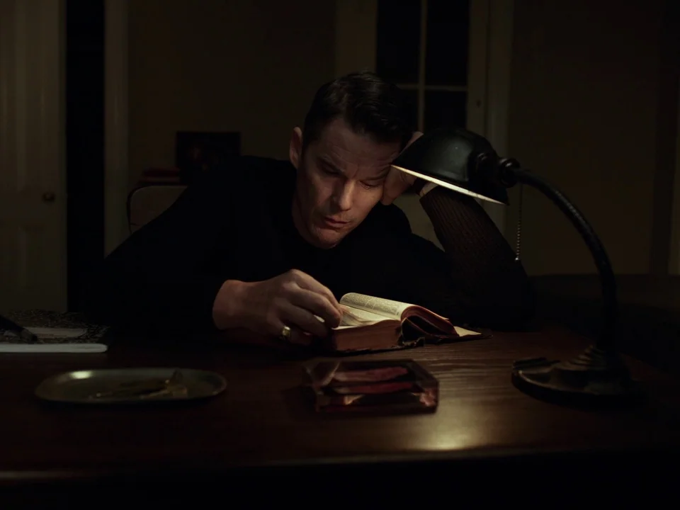 First Reformed (2017) movie still: medium — A dimly lit room contains a wooden desk with a lamp and a book. A man sits at the desk reading the book…