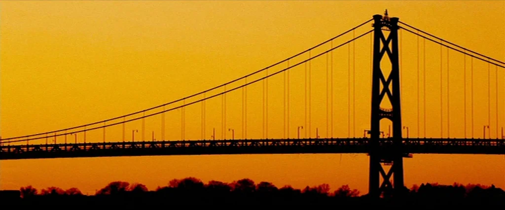 Hobo With A Shotgun (2011) movie still: establishing — A large suspension bridge spans the horizon against a vibrant orange sunset sky. The bridge…