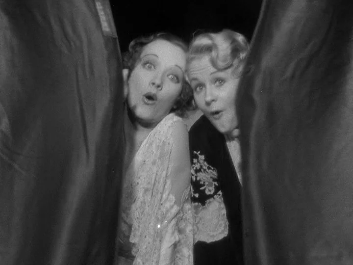 42nd Street (1933) movie still: medium — Two women peek through a narrow opening between two heavy dark curtains indoors. They have wide-eyed…