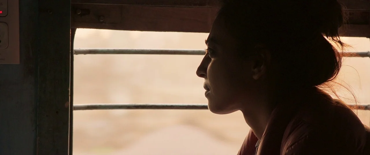 The Wedding Guest (2018) movie still: closeup — A woman sits by a window inside a train carriage with metal bars visible. She looks out at the…