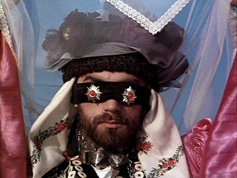 Ashik Kerib (1988) movie still: closeup — An open landscape provides a clear blue sky background for a costumed individual. The person wears an…