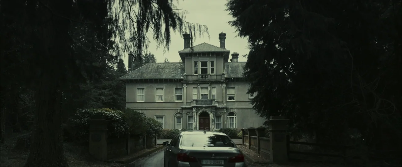 The Cellar (2022) movie still: wide — A large stone mansion with multiple windows stands behind a short wall and trees. A grey sedan is parked on…