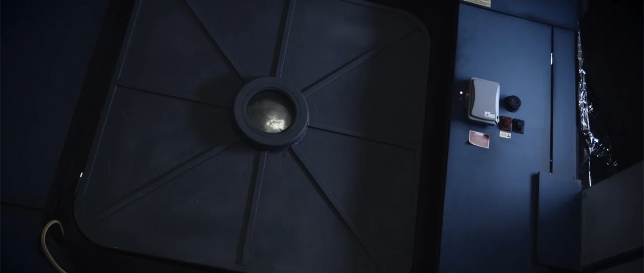 Astronaut: The Last Push (2012) movie still: medium — A dark industrial corridor features a heavy metallic door with a small central porthole. The…