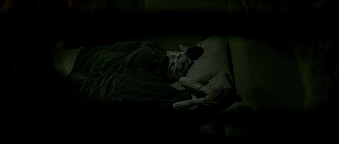 Dark Water (2005) movie still: medium — A dark bedroom shows two figures lying in a bed under grey blankets. One person is positioned next to a…