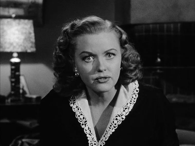 In a Lonely Place (1950) movie still: medium — A dark indoor setting features a table lamp glowing behind a woman. The woman stares at the camera…
