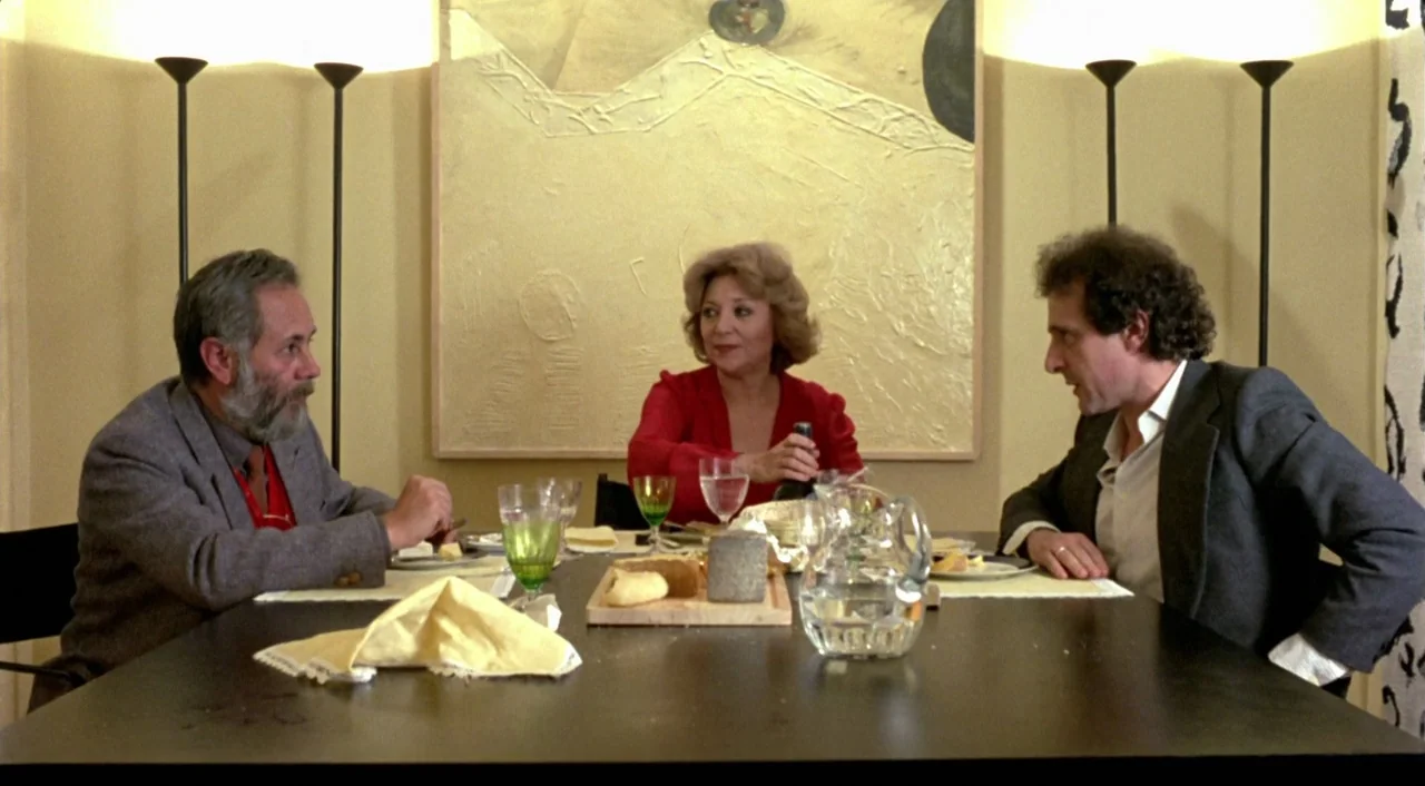 What Have I Done To Deserve This? (1984) movie still: medium — A dining table is set with bread and glasses in a beige-toned room. Three adults sit…