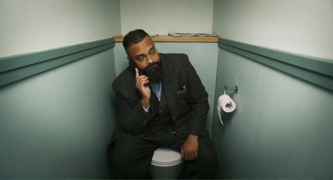 How to Date Billy Walsh (2024) movie still: medium — A narrow toilet cubicle with muted teal-painted walls and a wooden trim shelf. A man in a dark…