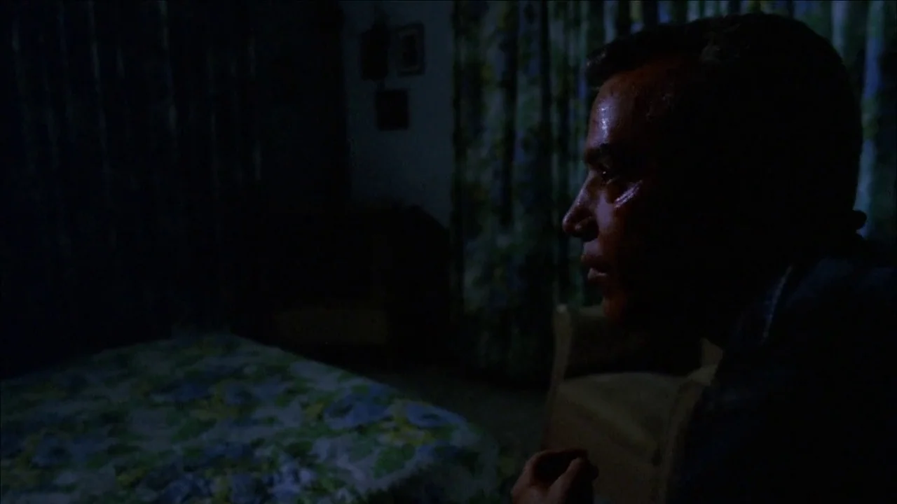 The Slumber Party Massacre (1982) movie still: medium — A darkened room features a patterned floral quilt on a bed and curtains. A man sits in…