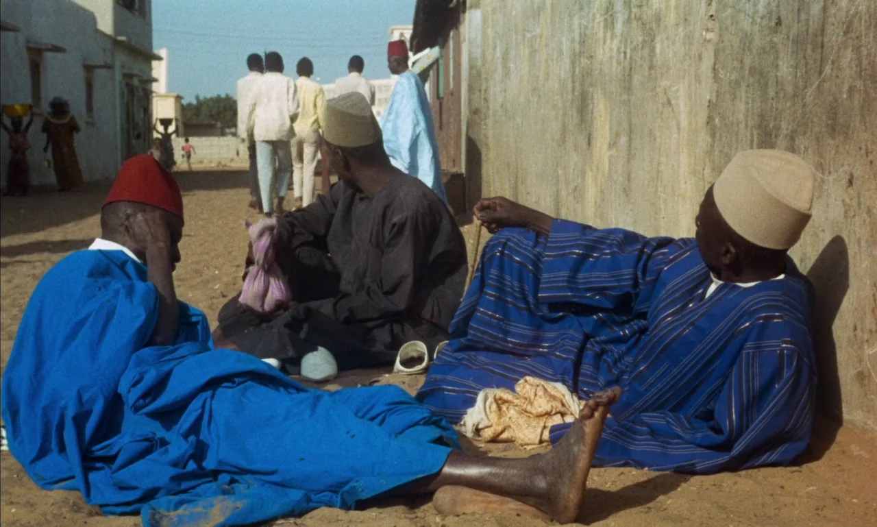 Mandabi (1968) movie still: medium — Three men in traditional West African robes sit on the sandy ground beside a wall. Other figures walk along a…