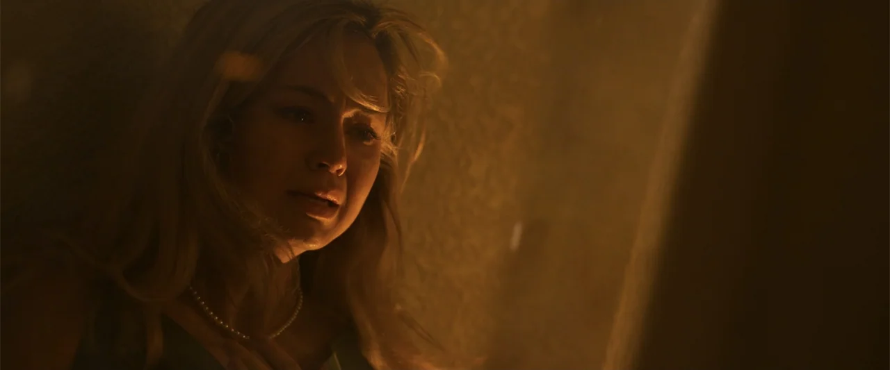 Final Destination Bloodlines (2025) movie still: closeup — A dark indoor space is illuminated by a single warm light source. A blonde woman in her…