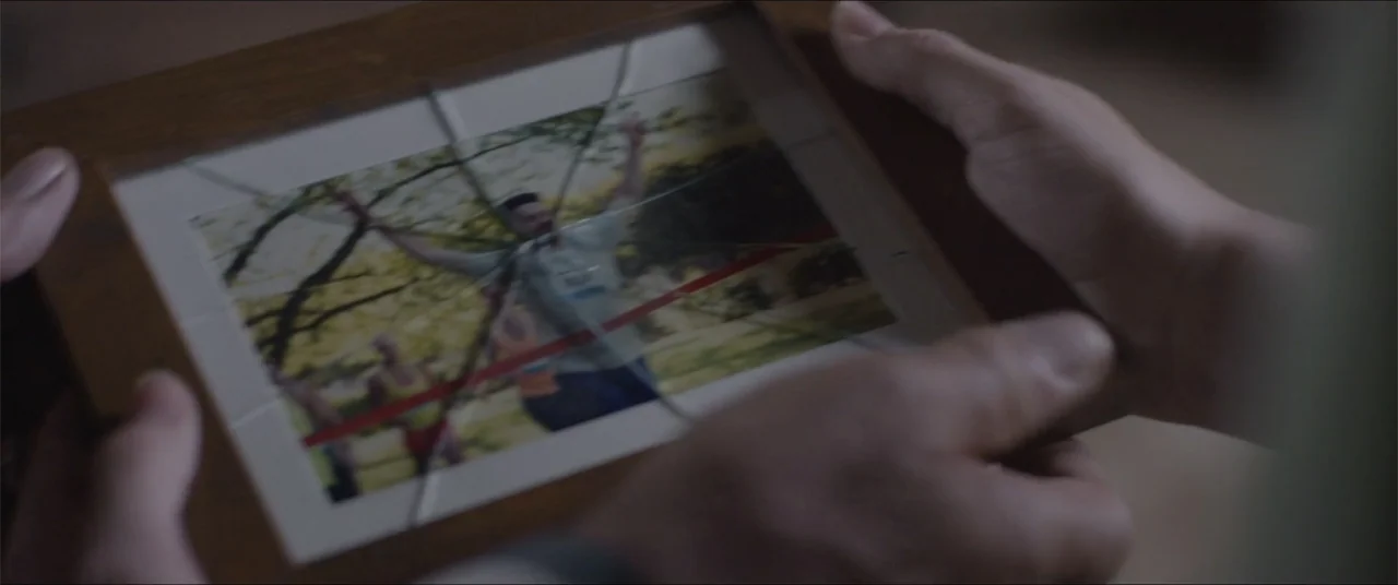 Captive (2020) movie still: closeup — A close view shows two hands holding a wooden picture frame with cracked glass. The photo inside depicts a…