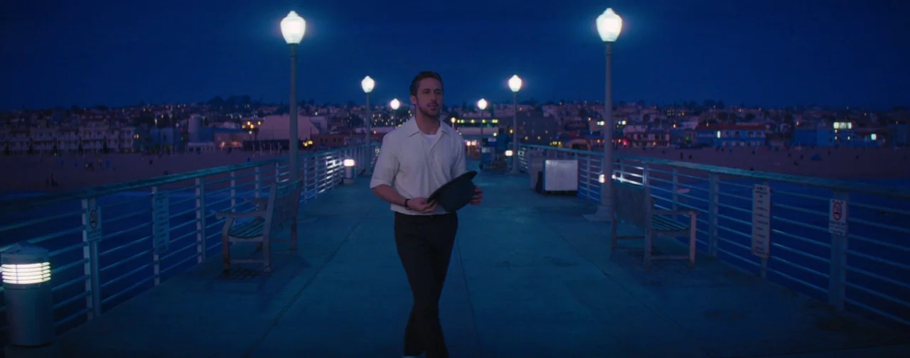 La La Land (2016) movie still: fullbody — A wooden pier stretches out into the dark night, illuminated by evenly spaced streetlamps. A man in a…